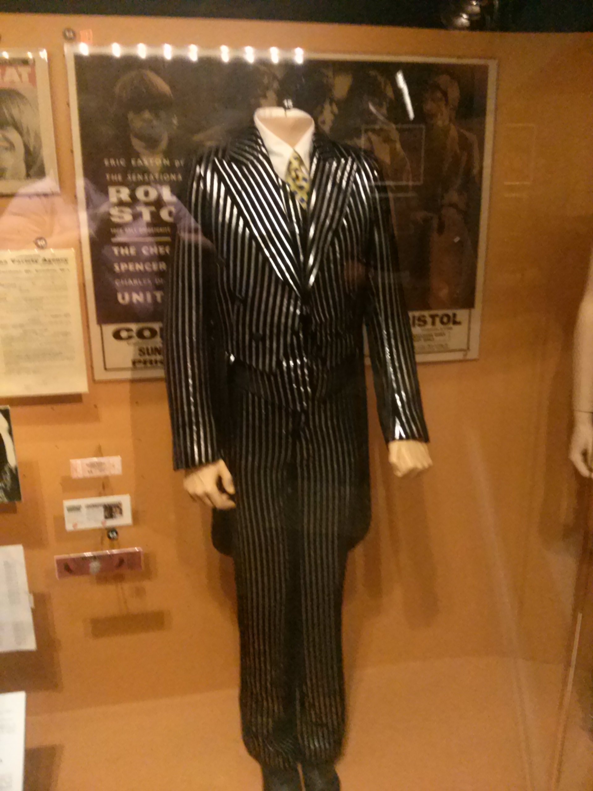Photograph: The Rolling Stones exhibit with a headless mannequin with black and silver striped pants suit jacket with white shirt underneath jacket and a yellow tie with black spots. Behind mannequin are posters and other Rolling Stones memorabilia.